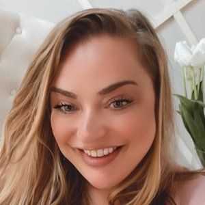 Woman with blonde hair smiles, indoors, next to white flowers and a patterned backdrop.