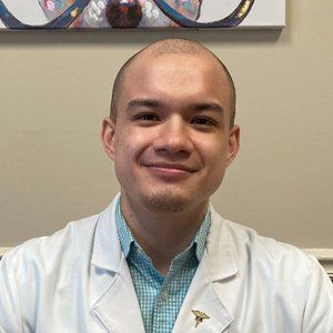 Man wearing a white coat and light blue shirt smiles at the camera; art in the background.