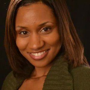 Woman with brown hair smiles, wearing a green sweater against a black background.
