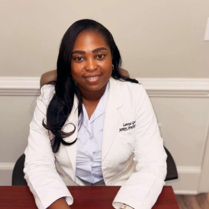 Woman in white lab coat sitting at a desk, smiling.