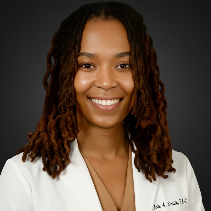 Smiling woman in a white coat, with brown dreadlocks. Name tag visible. Neutral background.