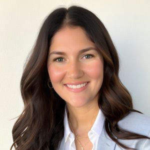 Woman with long brown hair, smiling, wearing a white shirt and light blue blazer.