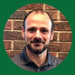 Man with a beard in a gray polo shirt in front of a brick wall, inside a green circle.