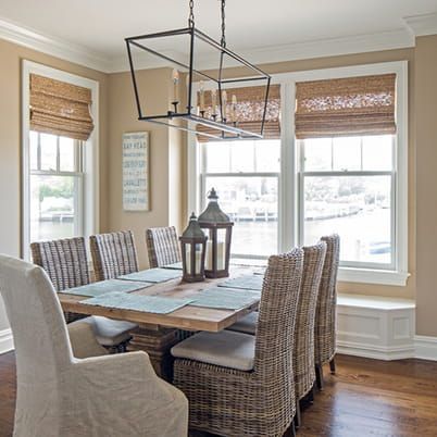 Dining room with wicker chairs, wood table, and large windows with woven shades