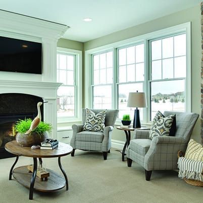 Bright living room with white fireplace, plaid chairs, large windows, and a round coffee table with decor