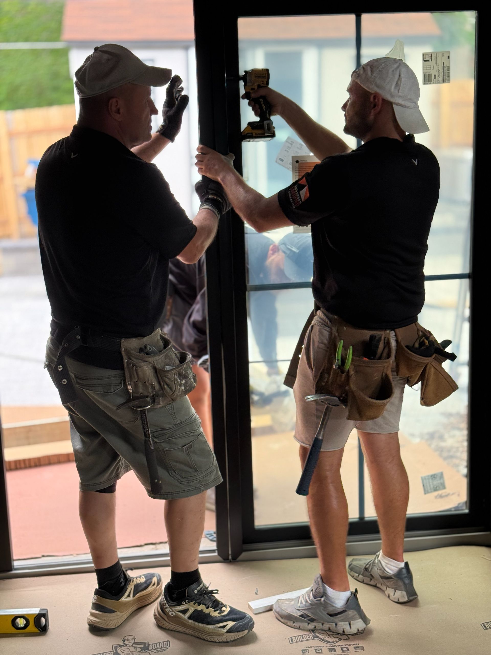Two workers installing a sliding glass door inside a bright room, using tools and scaffolding belts.