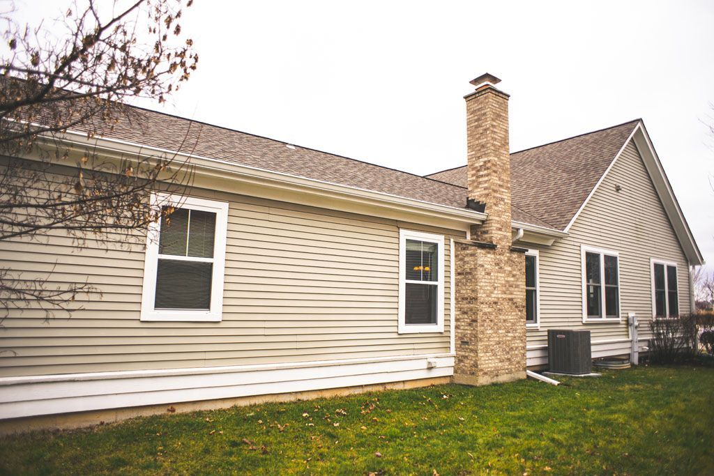 Single-story beige house with a stone chimney, white-trimmed windows, and a grassy yard