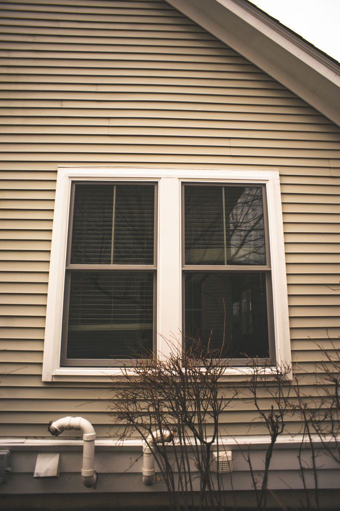 White-trimmed window on a beige house exterior with pipes and bare shrubs below.
