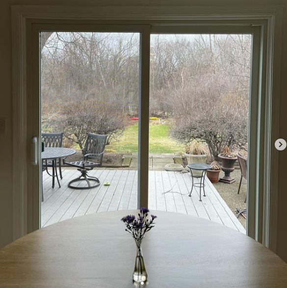 Dining table by a sliding glass door overlooking a deck, patio furniture, and a wooded backyard.