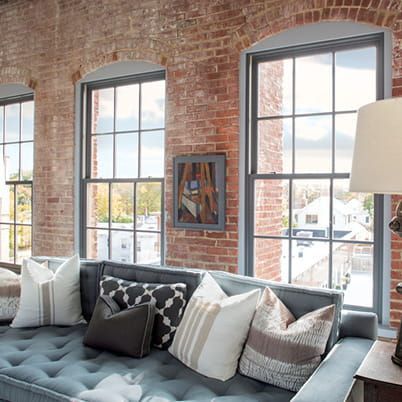 Cozy loft living room with exposed brick, large windows, and a gray sectional sofa with pillows and a lamp