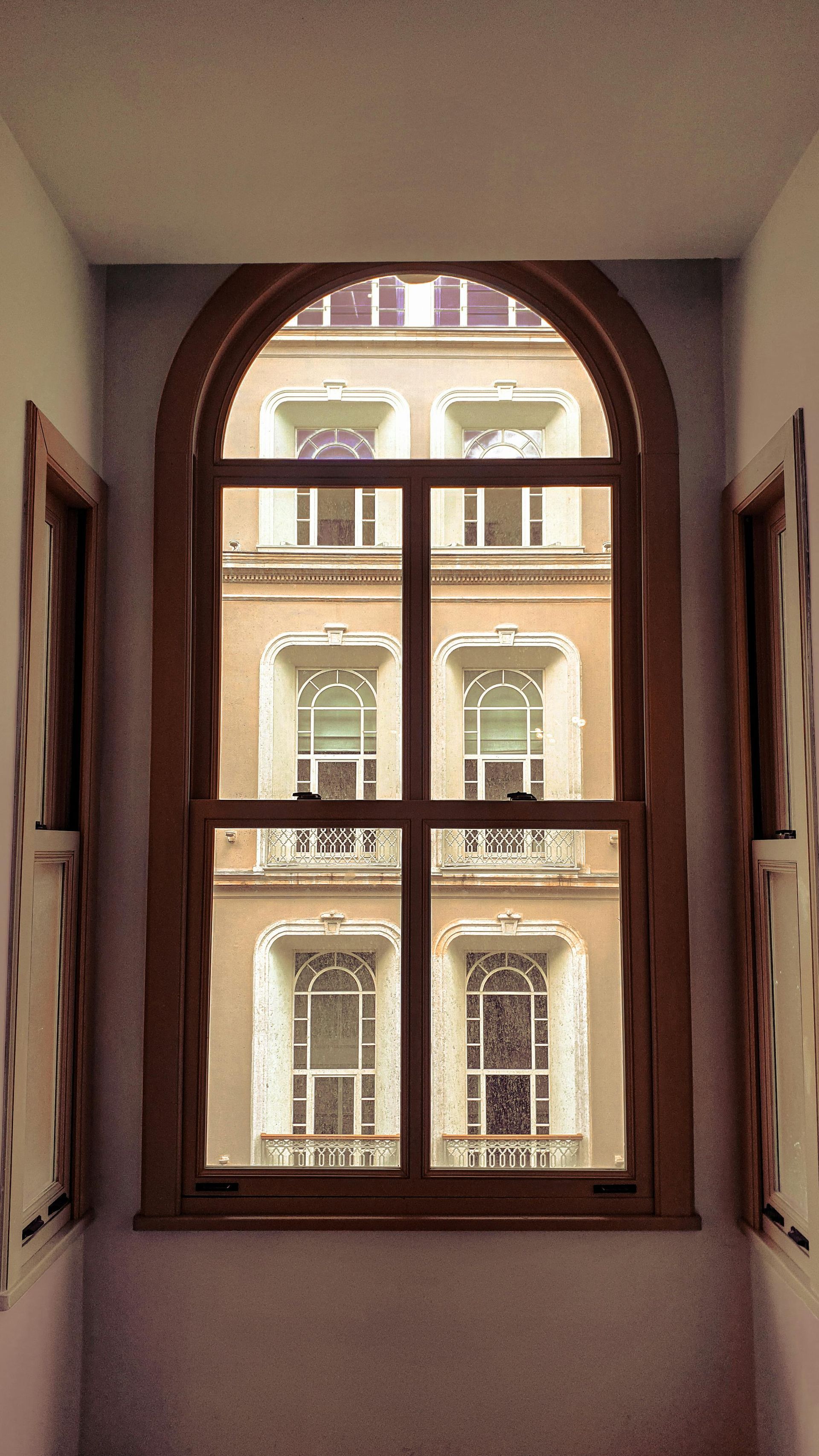 Arched window with wooden frame overlooking a beige building with ornate white-trimmed windows.