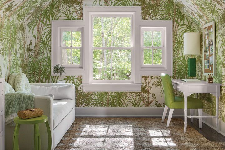 Bright living room with leafy wallpaper, white sofa, green chairs, desk, and large windows overlooking trees