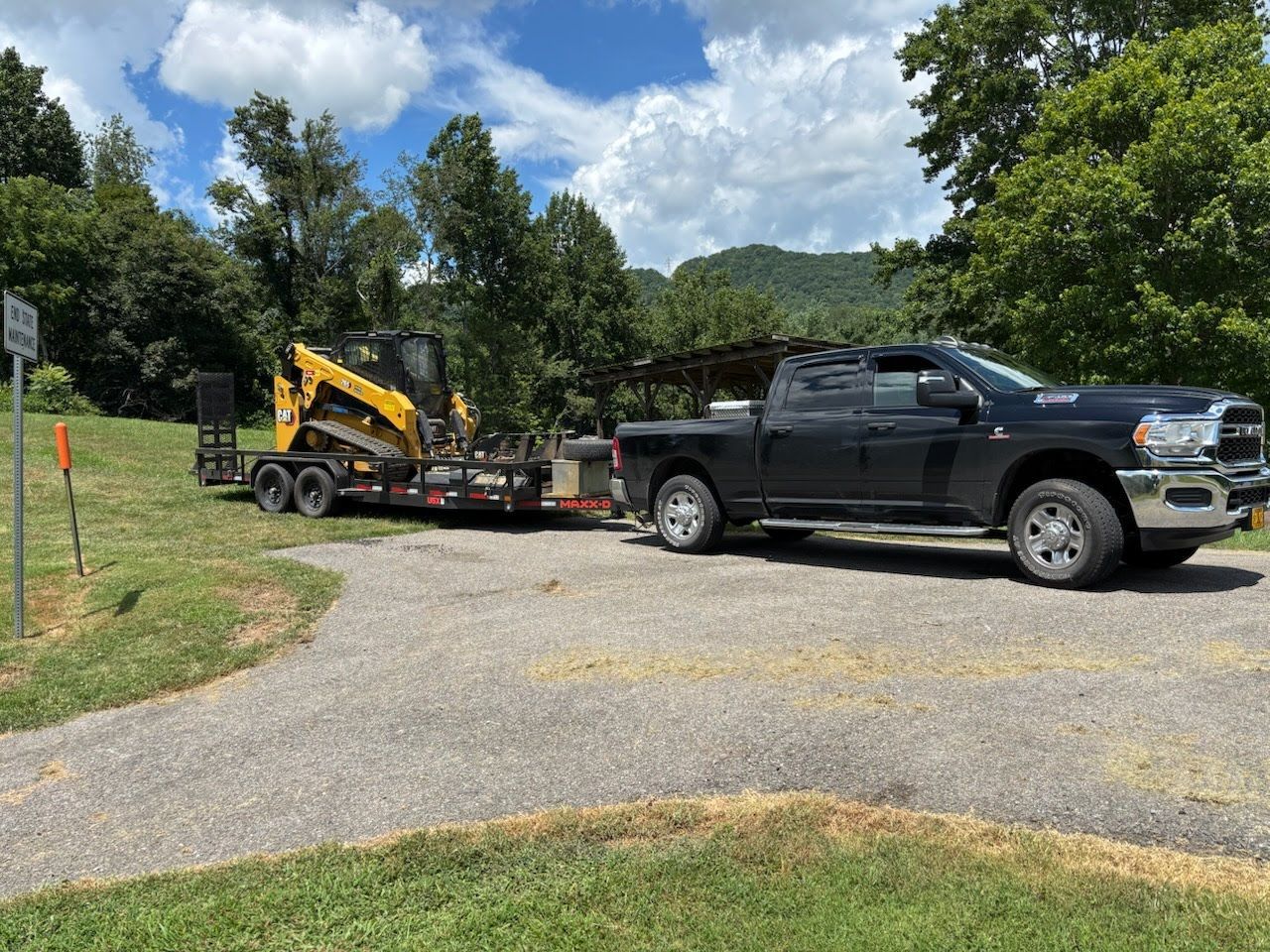 A black pickup truck towing a flatbed trailer with a yellow skid steer loader on a paved road near a grassy field.