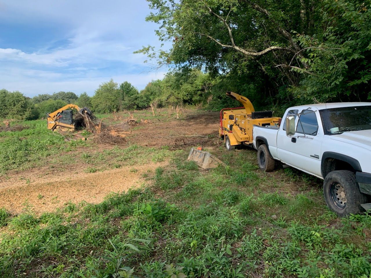 A white truck is hitched to a yellow wood chipper in a grassy field with land clearing equipment nearby.