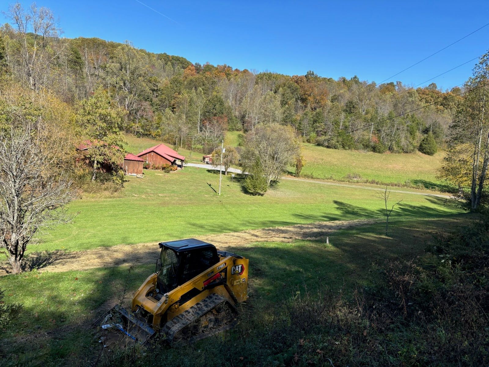A yellow skid steer loader sits on a grassy hill overlooking a rural landscape with red buildings and a forested mountain.