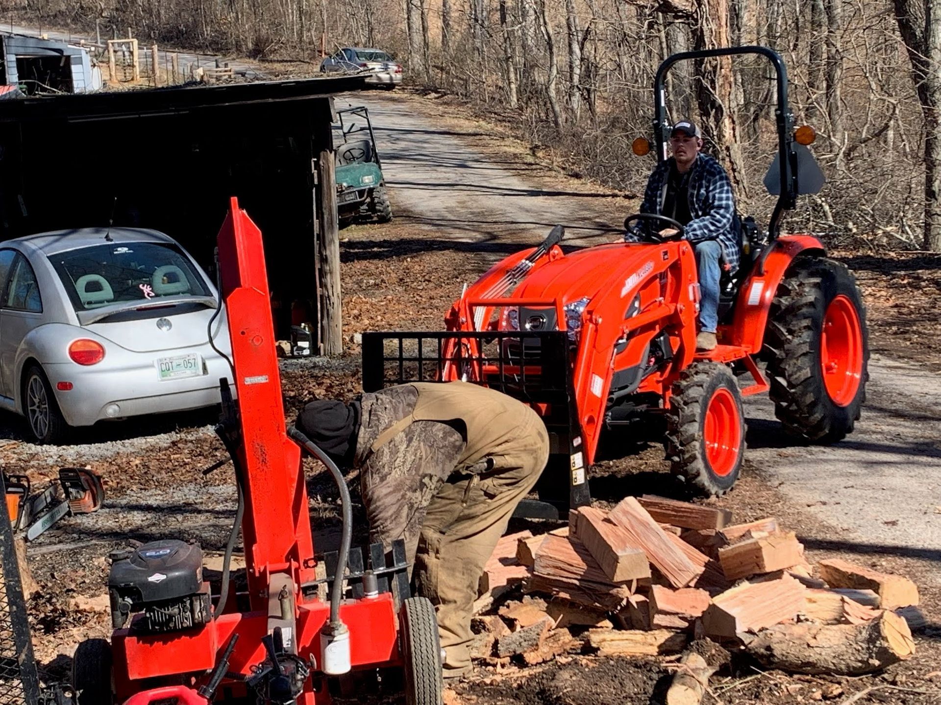 A person in camouflage coveralls works with a wood splitter while another person sits on a bright orange tractor nearby.