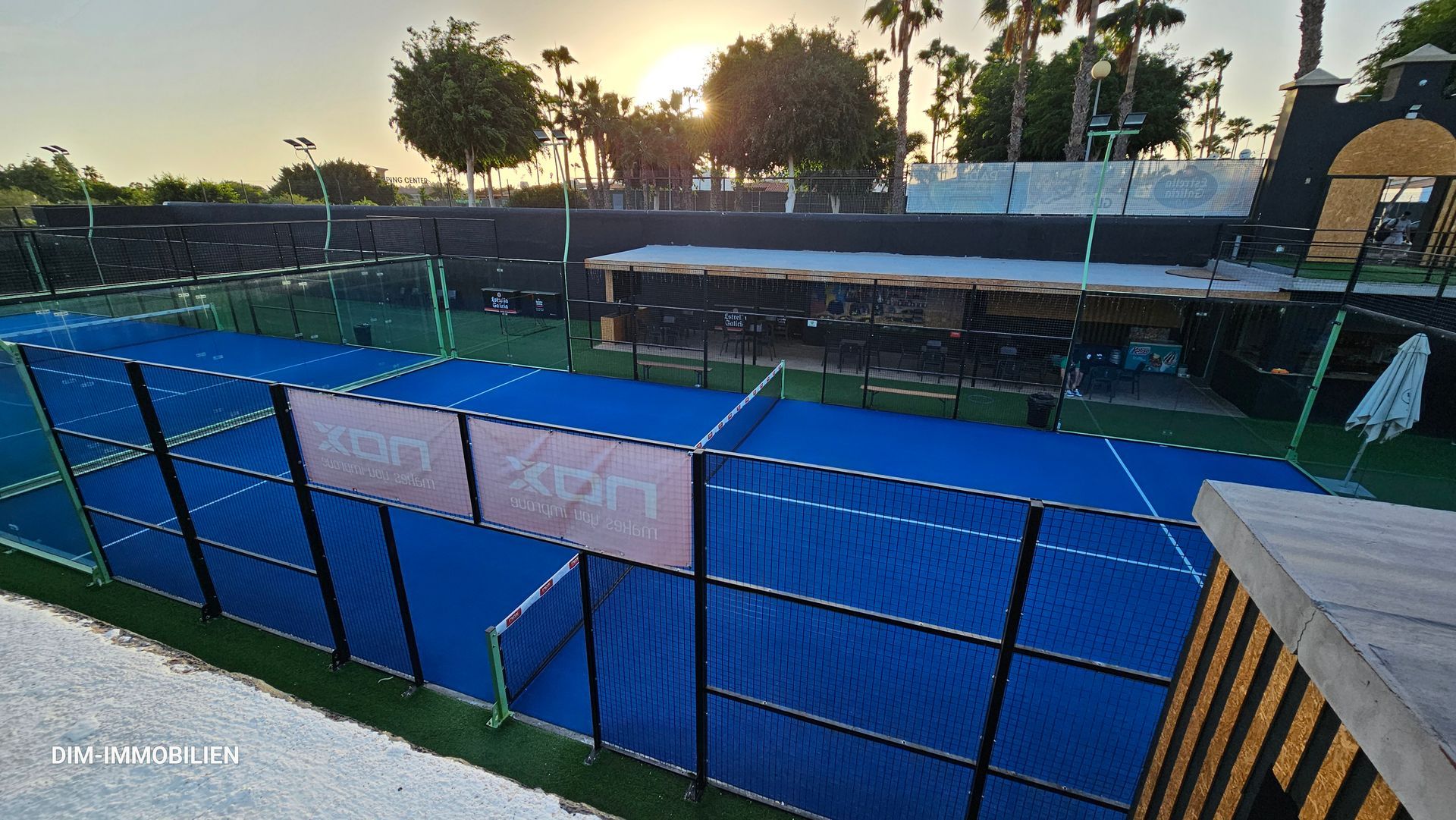 Ein Tennisplatz mit einem Zaun darum und der Sonne, die durch die Bäume scheint