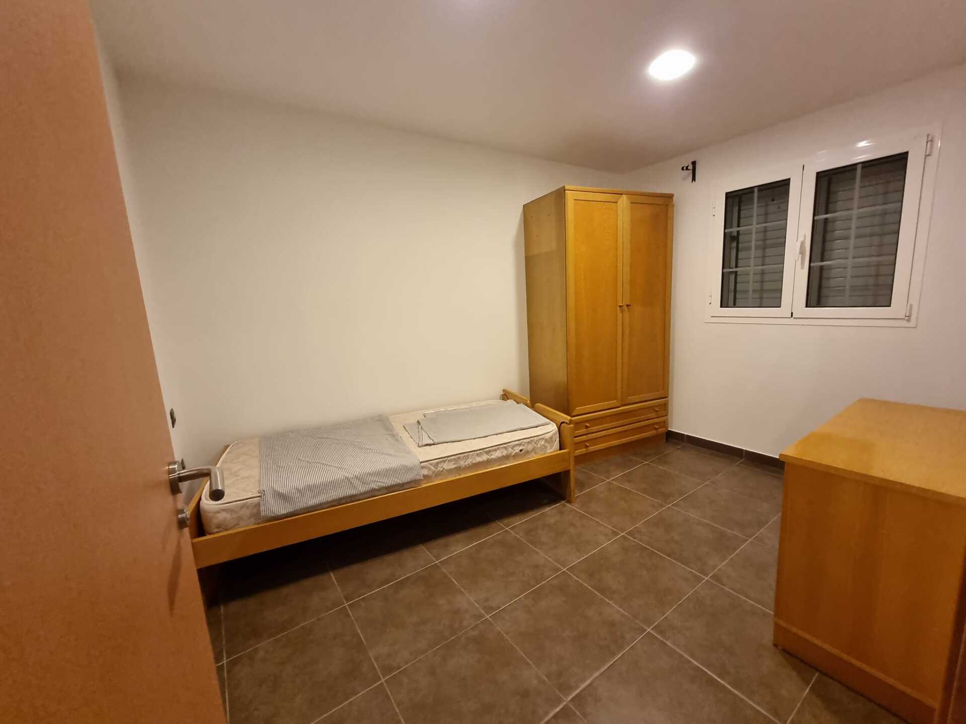 Ein Schlafzimmer mit Einzelbett, Kommode, Kleiderschrank und zwei Fenstern.