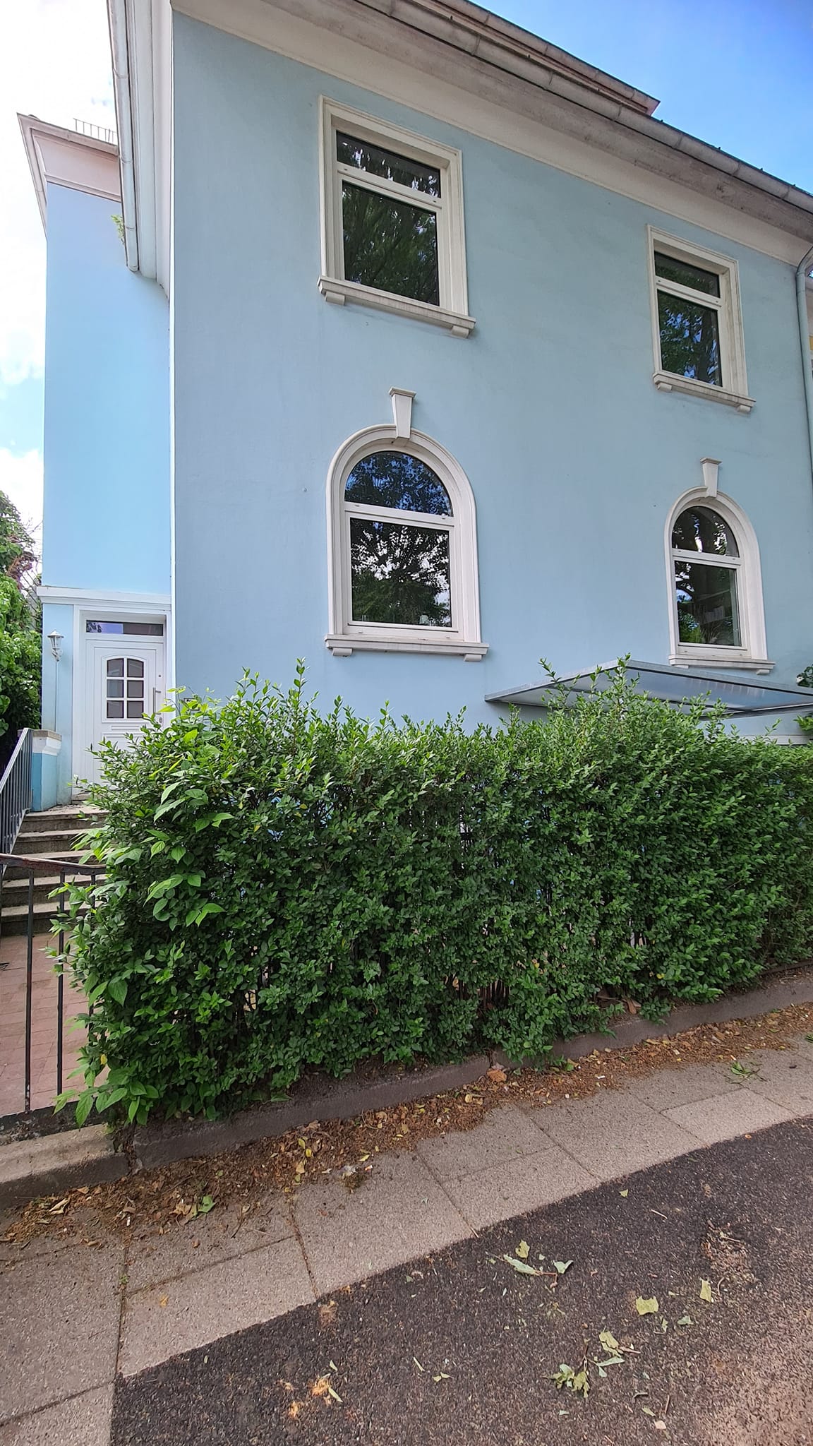 Ein blaues Gebäude mit einer Hecke davor und einer Treppe, die hinaufführt.