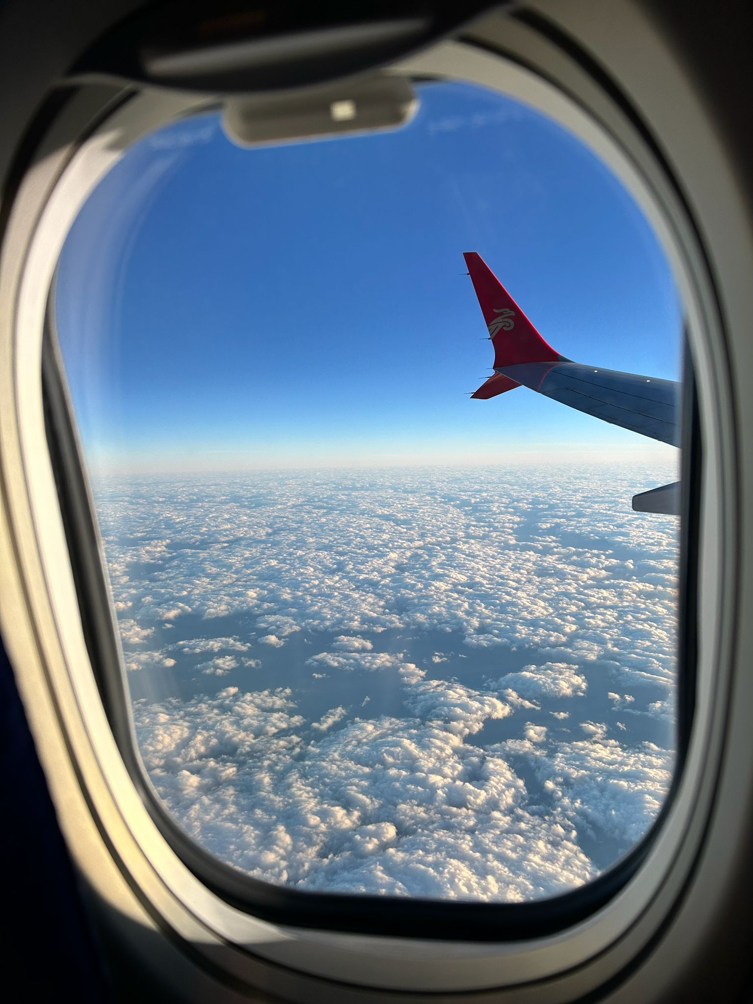 View from an airplane window over a sea of clouds and blue sky, with a red winglet visible.