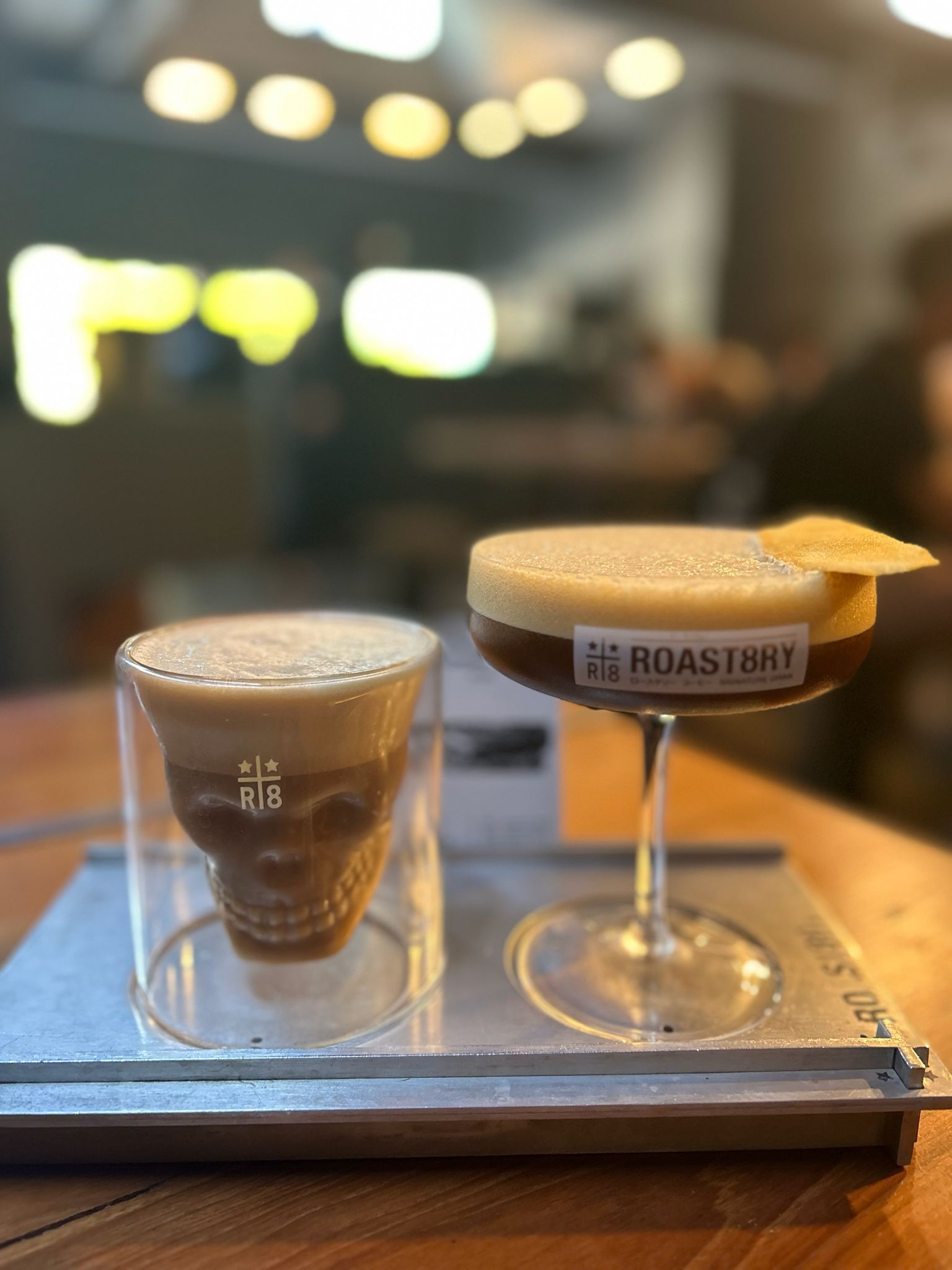 Two coffee cocktails on a tray: a clear glass and a stemmed coupe with foam and a citrus garnish.