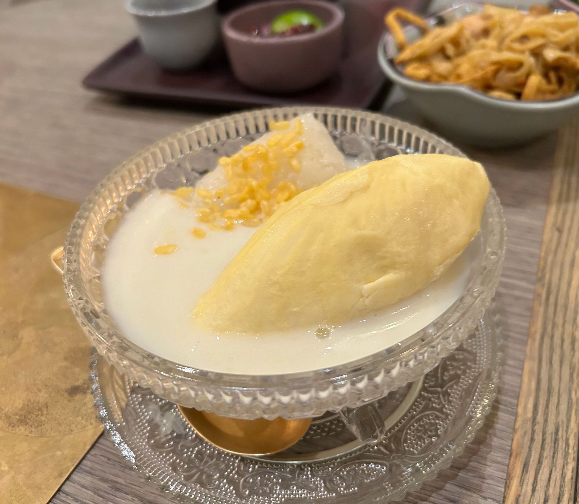 Thai mango sticky rice in a glass dish on a table, with bowls of dessert in the background