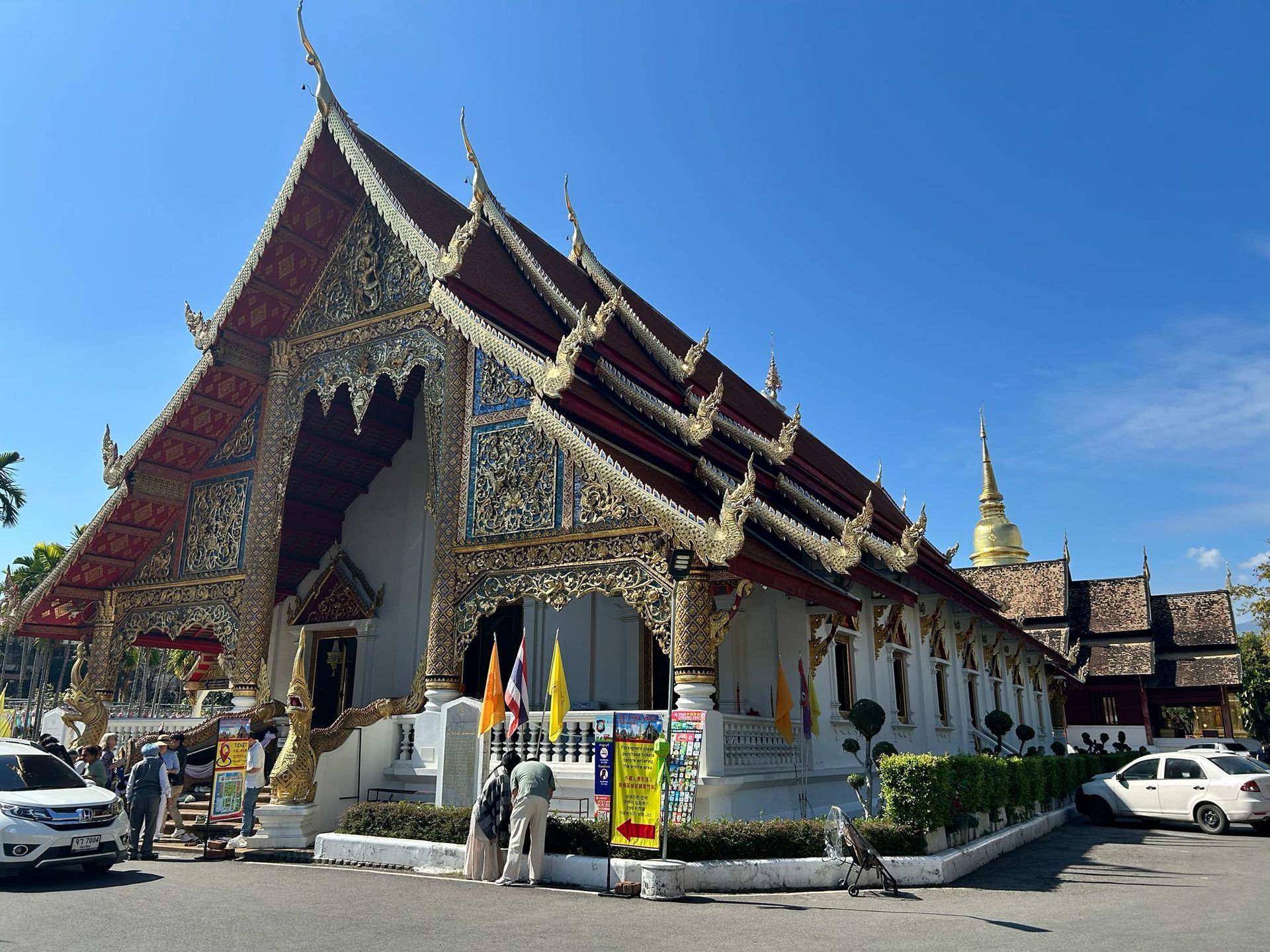 Ornate Thai temple with red-and-gold roof, white walls, and parked cars under a clear blue sky