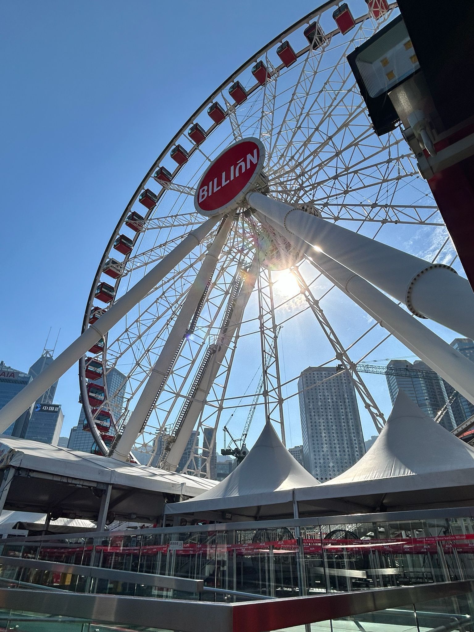 Ferris wheel above white tents in a bright city plaza, with sun glare and modern buildings nearby