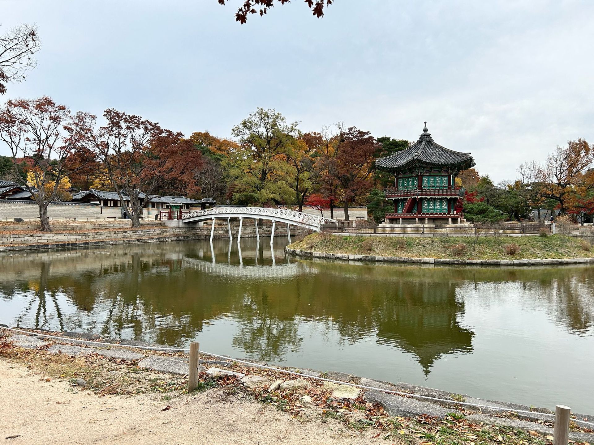 Traditional pavilion by a pond with autumn trees and their reflections under a cloudy sky
