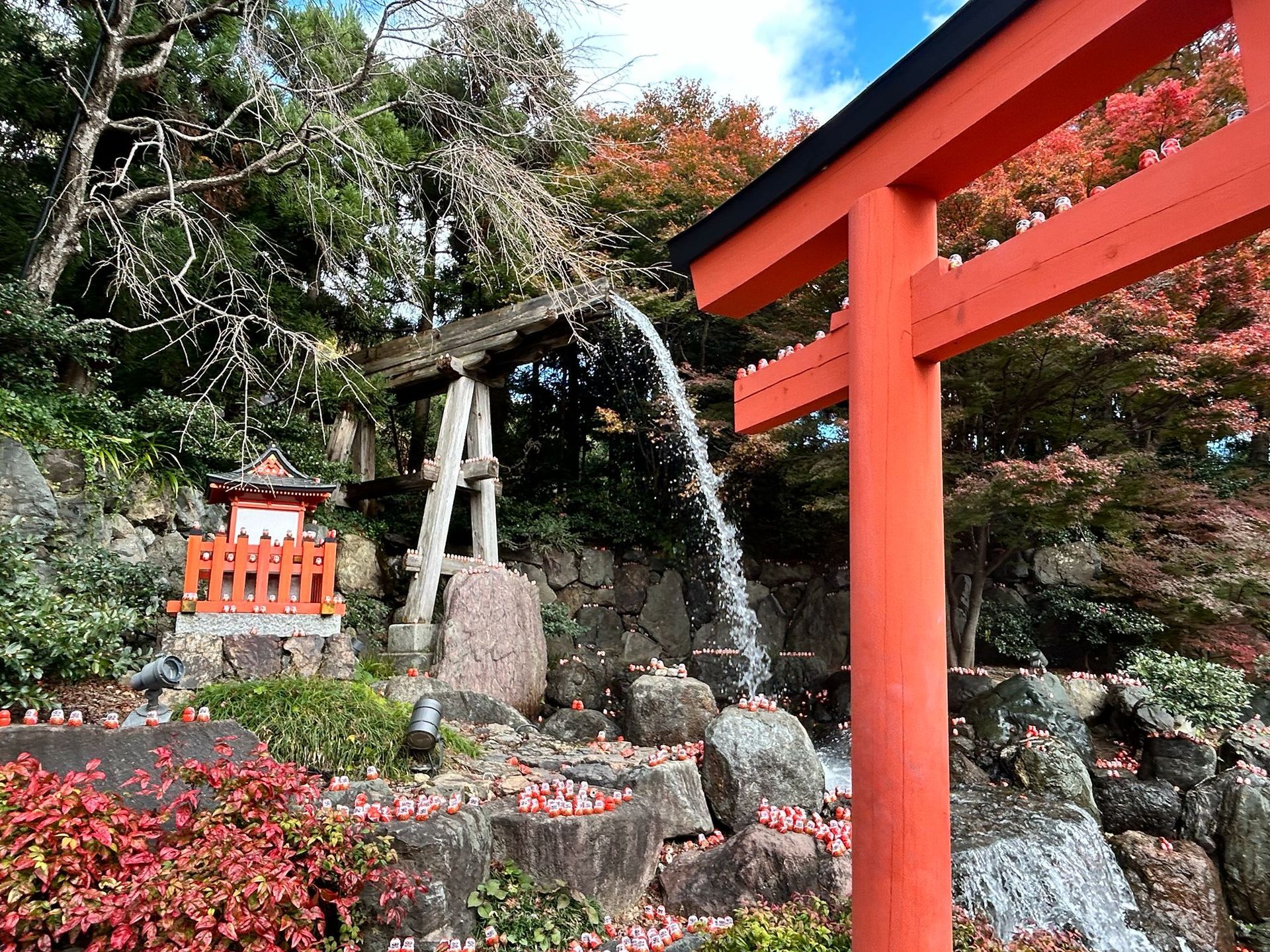 Red torii gate and stone shrine amid autumn foliage and rocks, with a small shrine in the background