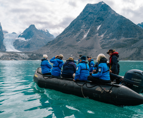 Six people in blue jackets ride a black inflatable boat on turquoise water near snowy mountains.