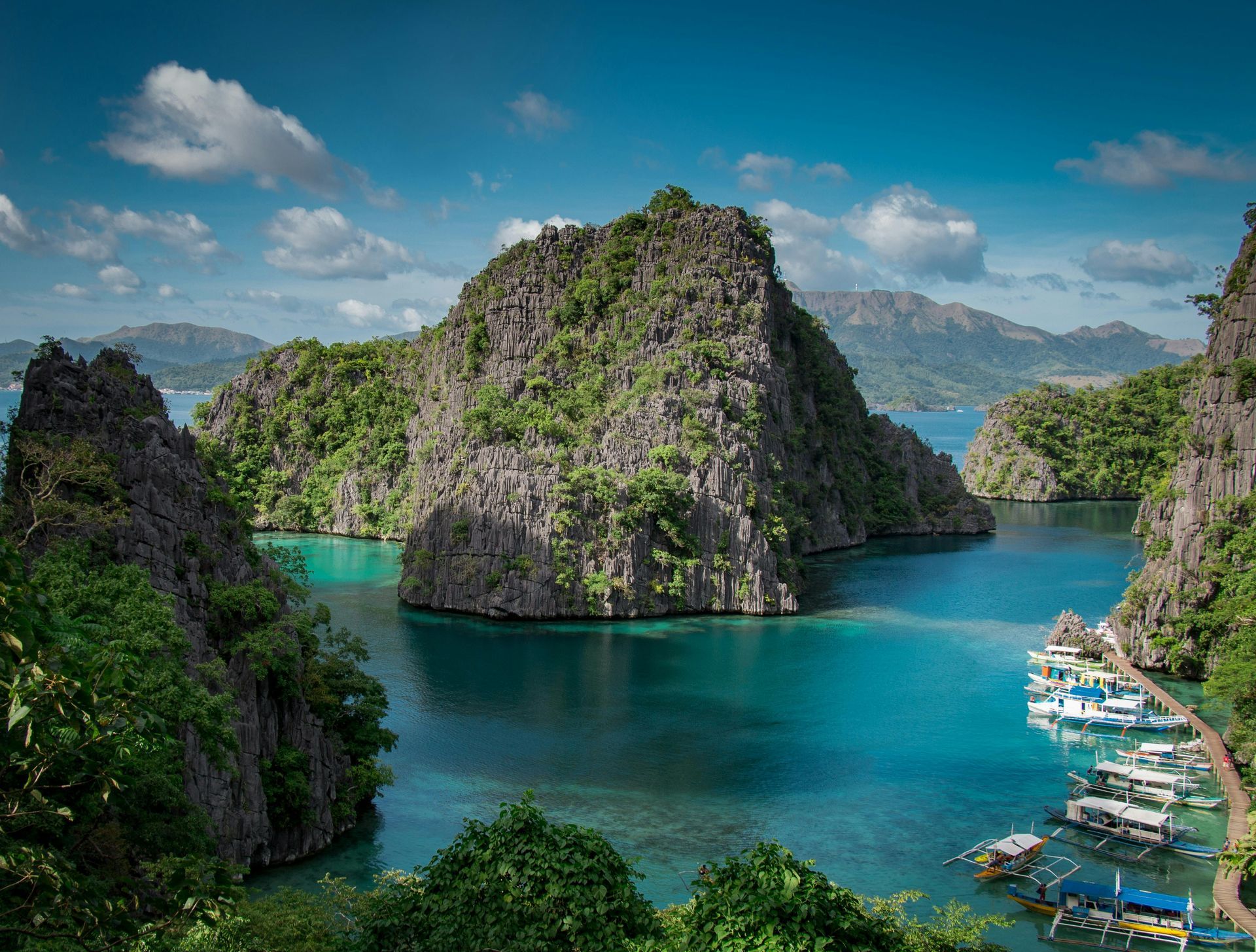 Turquoise bay with limestone cliffs, lush greenery, and boats under a bright blue sky