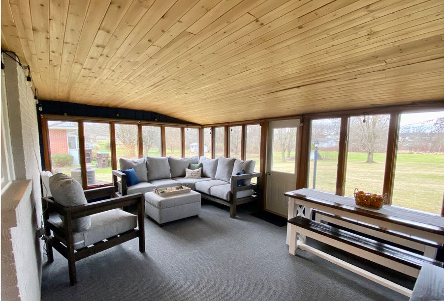 Bright sunroom with wood ceiling, gray sectional sofa, armchair, and large windows overlooking a grassy field