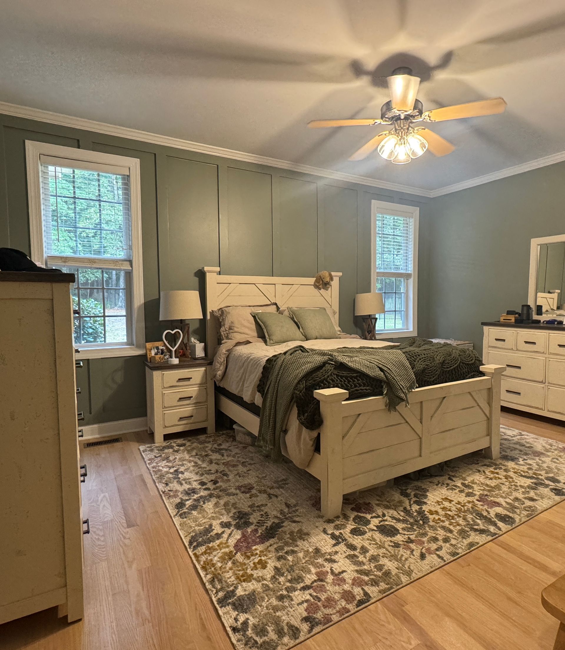 Cozy bedroom with a white bed, patterned rug, gray walls, windows, dresser, and ceiling fan.