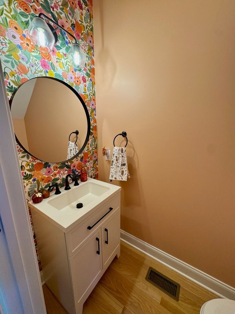 Small bathroom vanity with round mirror, floral accent wall, white sink, and tan walls.