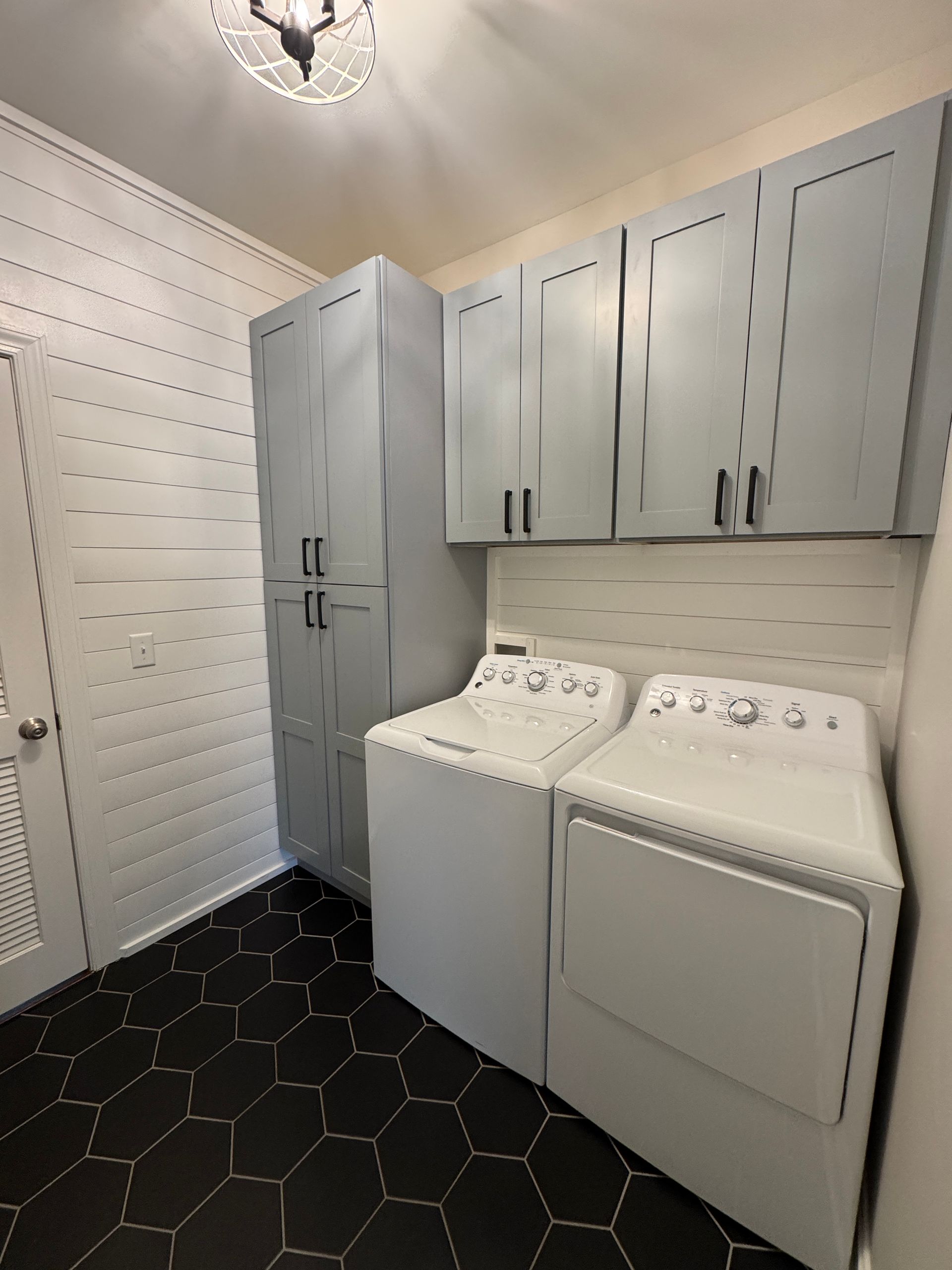 Small laundry room with gray cabinets, white washer and dryer, and black hex tile floor.