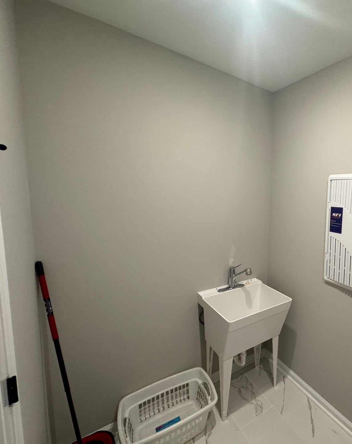 Small utility room with a white sink, laundry basket, and mop against light gray walls.