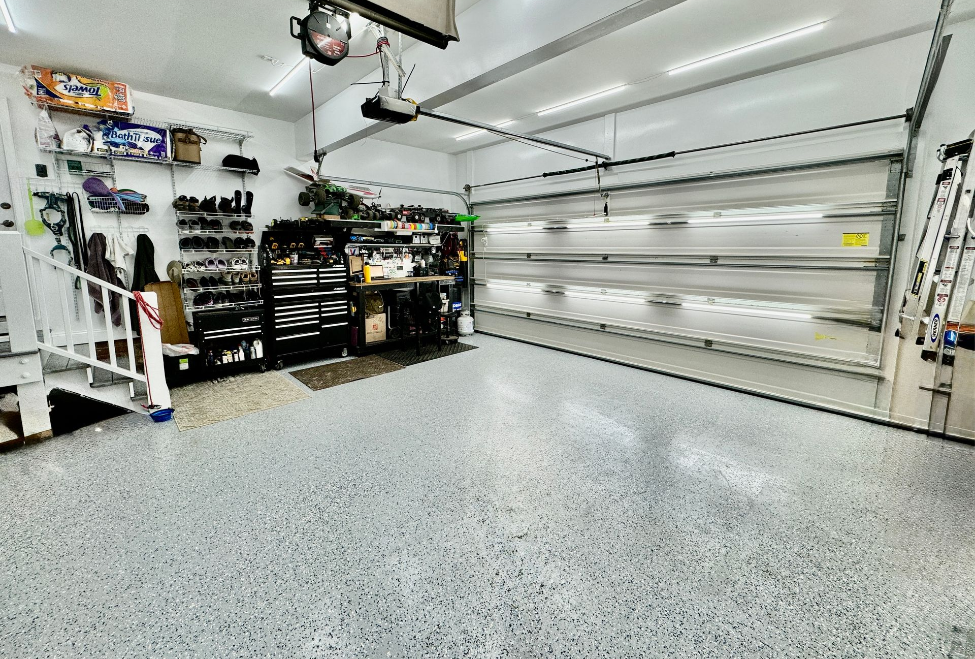 Bright garage with white walls, epoxy floor, shelves, tools, and an overhead door partially open