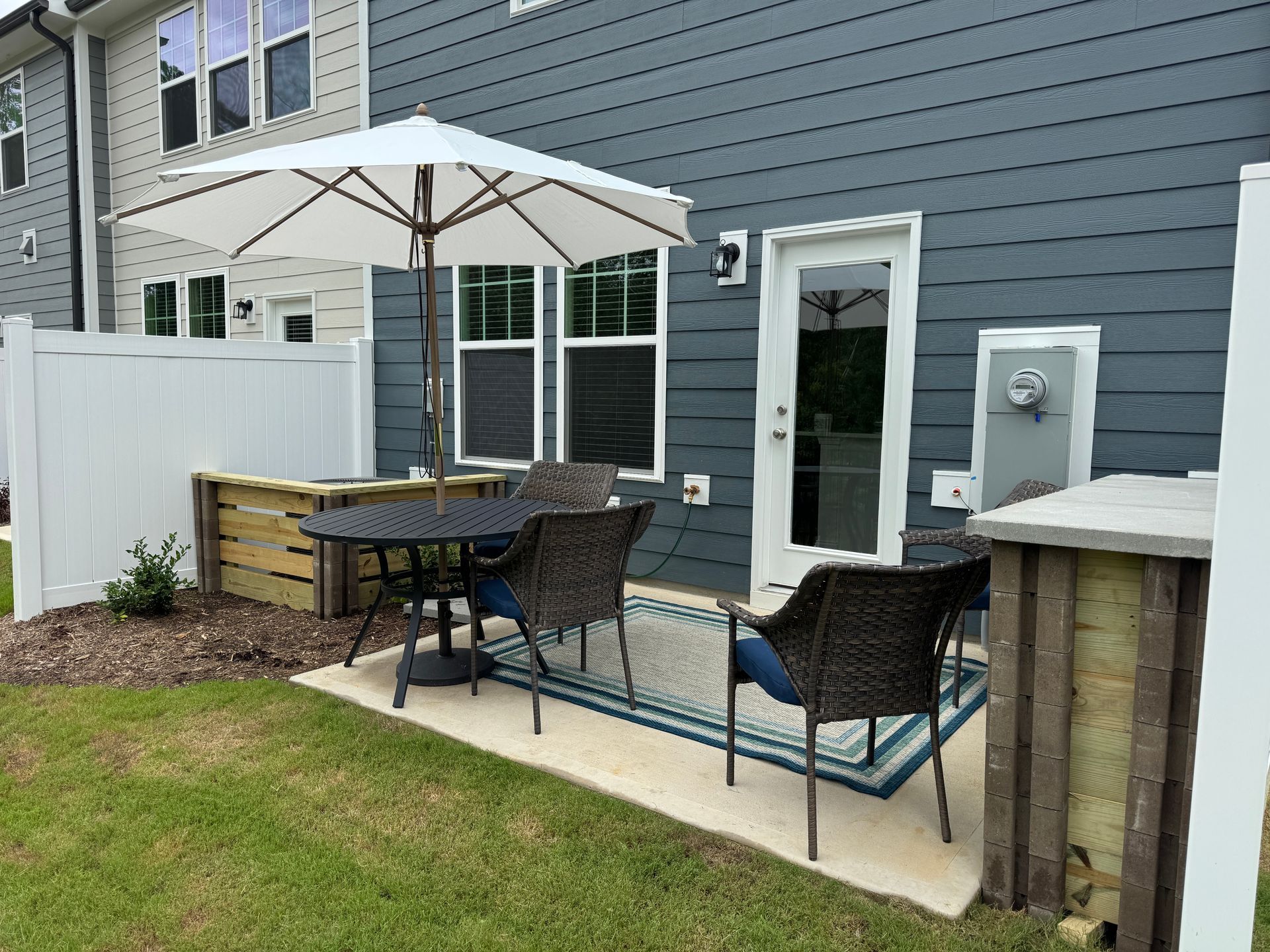 Patio with umbrella, table, chairs, and potted plants beside a gray house and white fence
