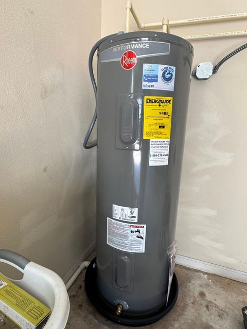 A grey Rheem electric water heater stands in a plastic drain pan on a floor next to a white wall in a residential setting.