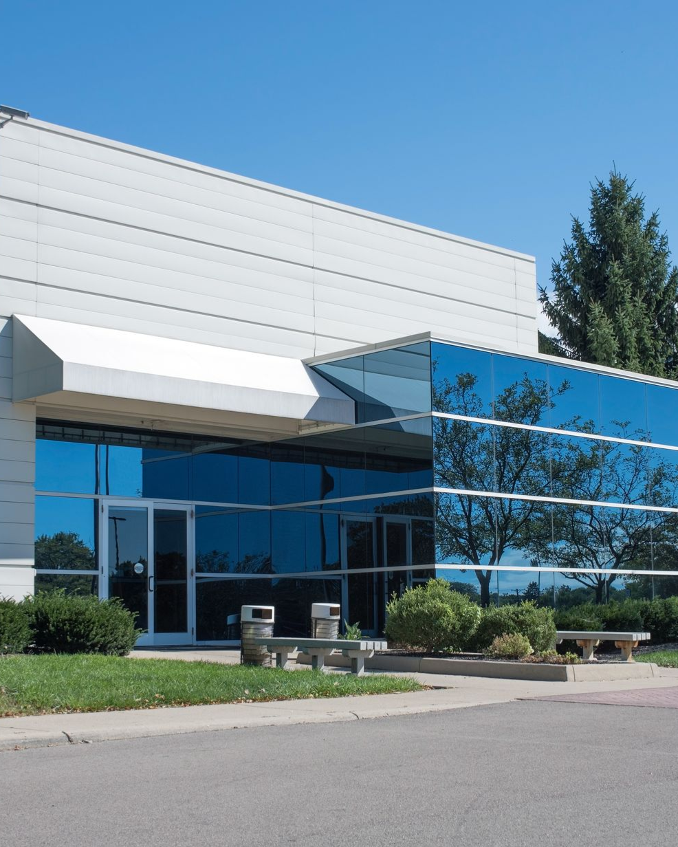 Modern building with reflective blue glass windows, entrance with canopy, and surrounding greenery.