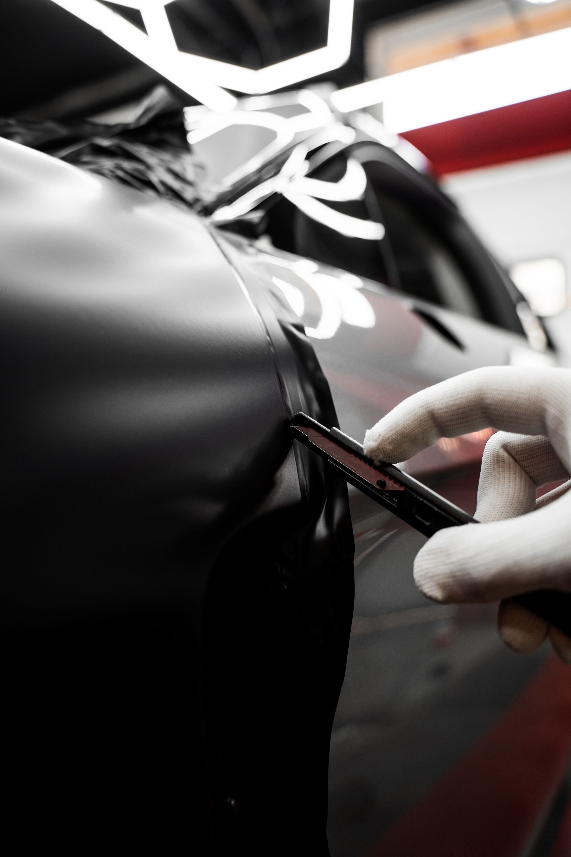 Person wearing a glove cuts black vinyl wrap on a car door with a blade in a garage.