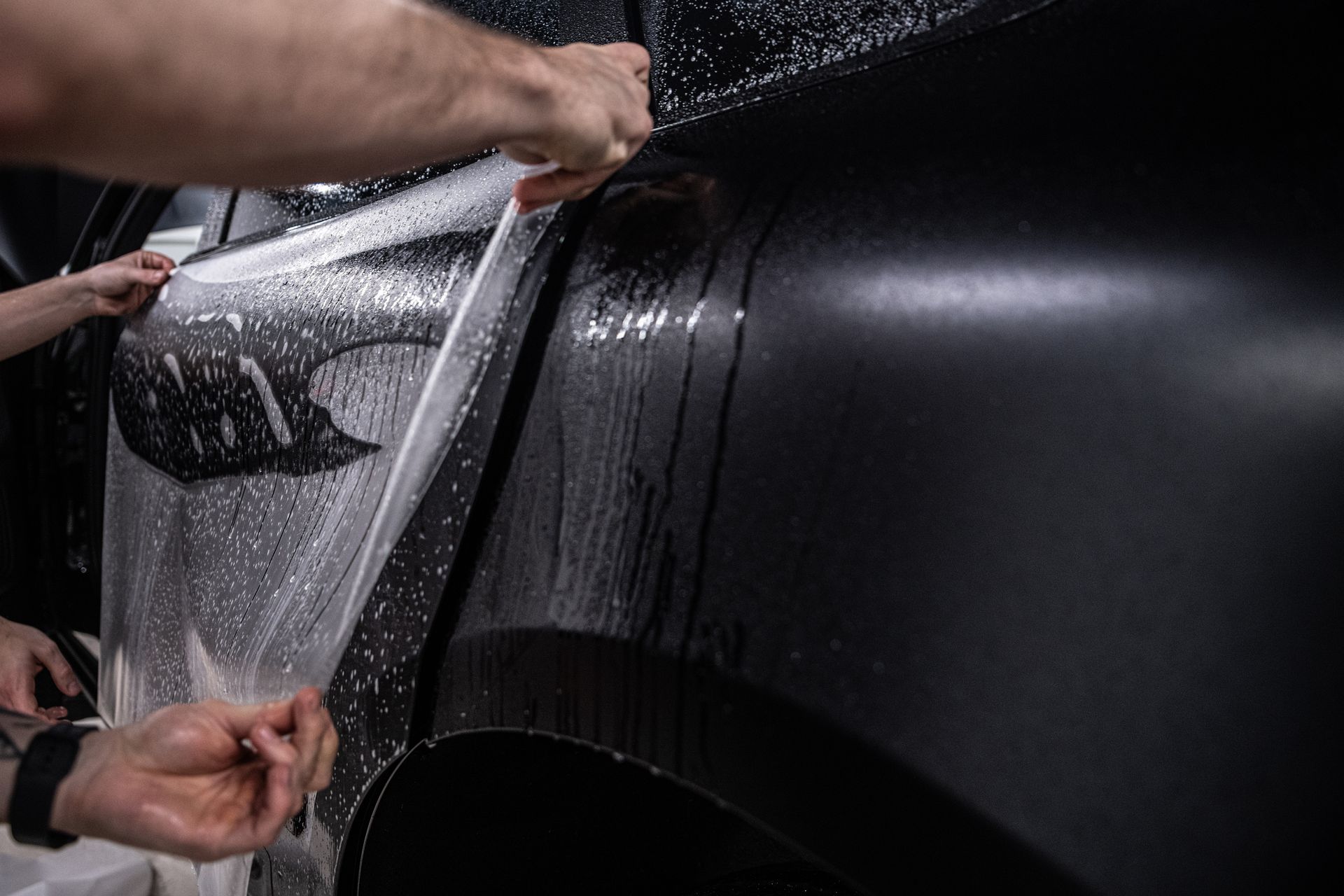 Hands applying wet protective film to a black car's side panel.