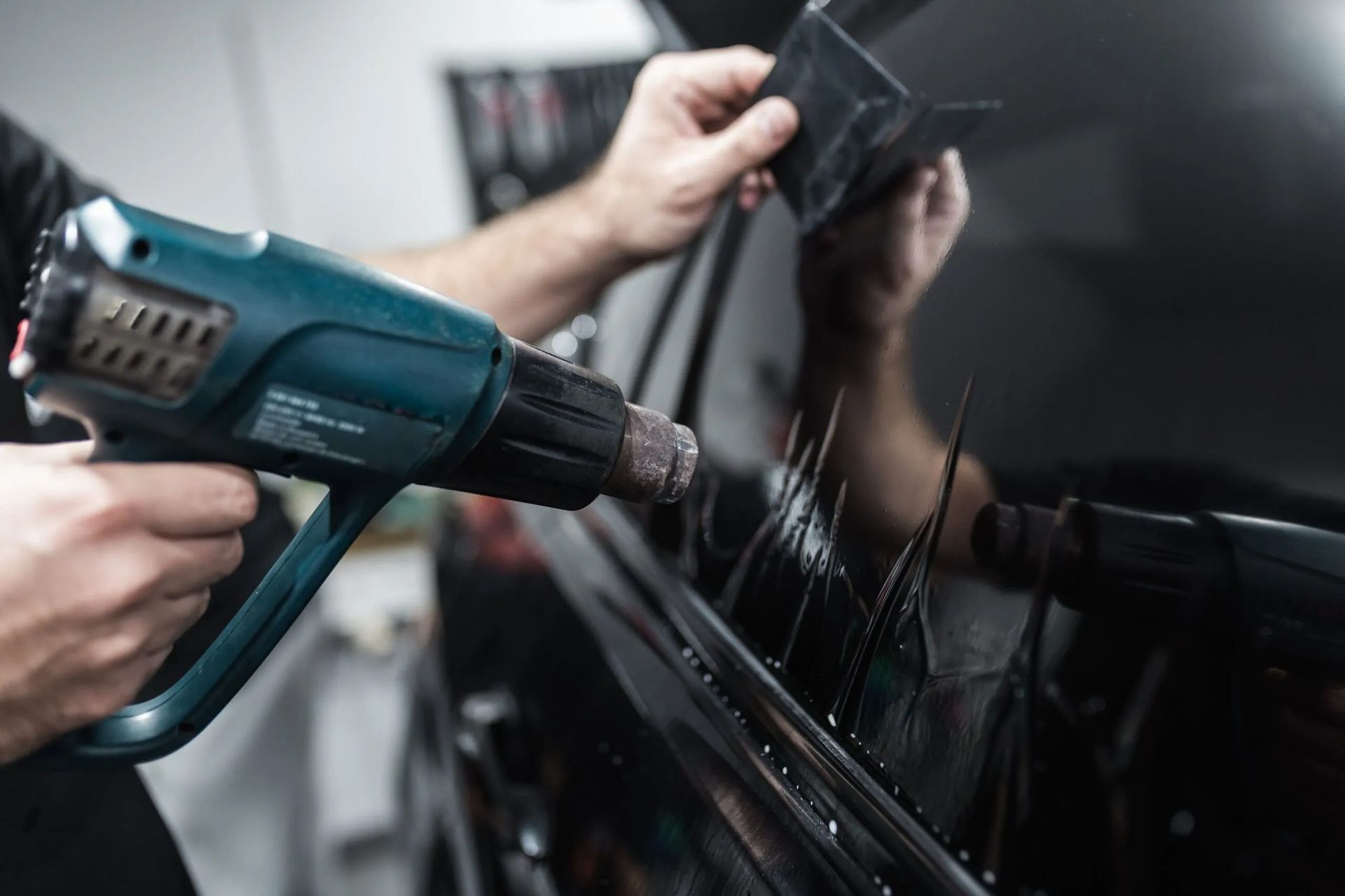 Person using a heat gun and squeegee to apply tinted film to a car window.