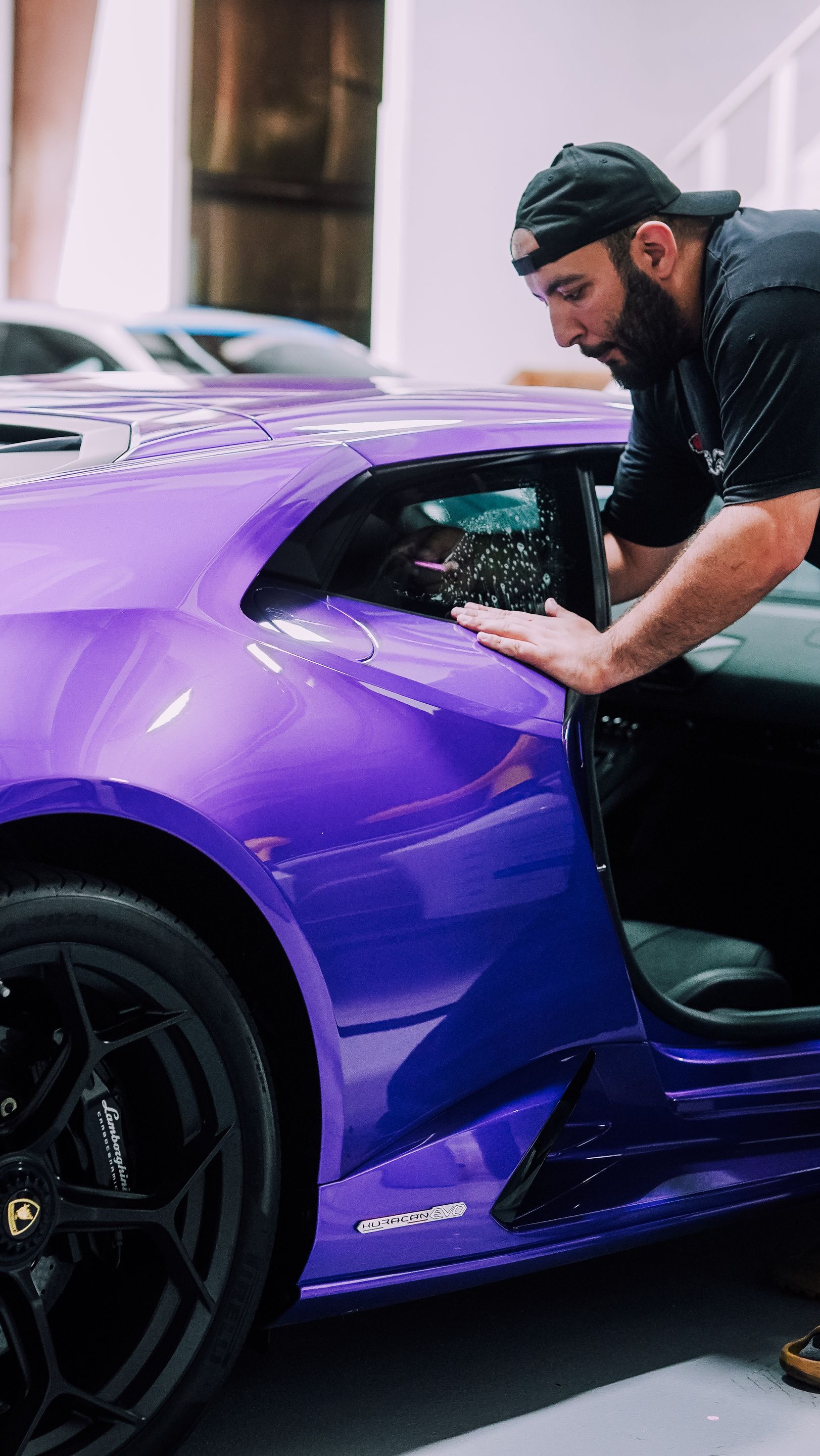 Man looking into a purple Lamborghini. He's wearing a black shirt and hat. The car is indoors.