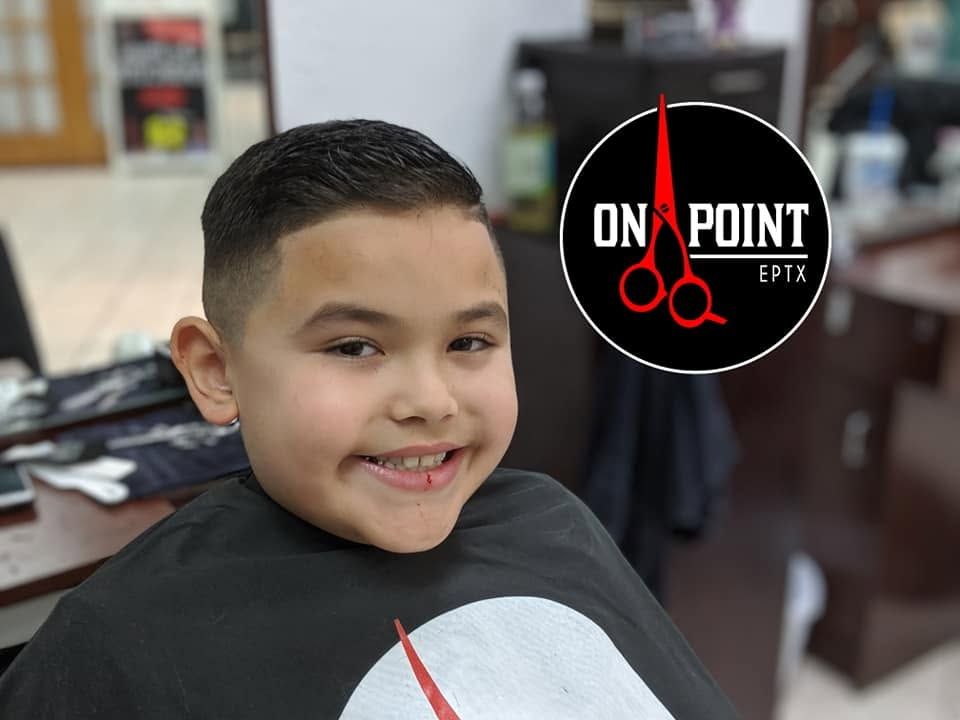 Boy smiling in a barber chair, wearing a black cape, with a scissors logo in the background