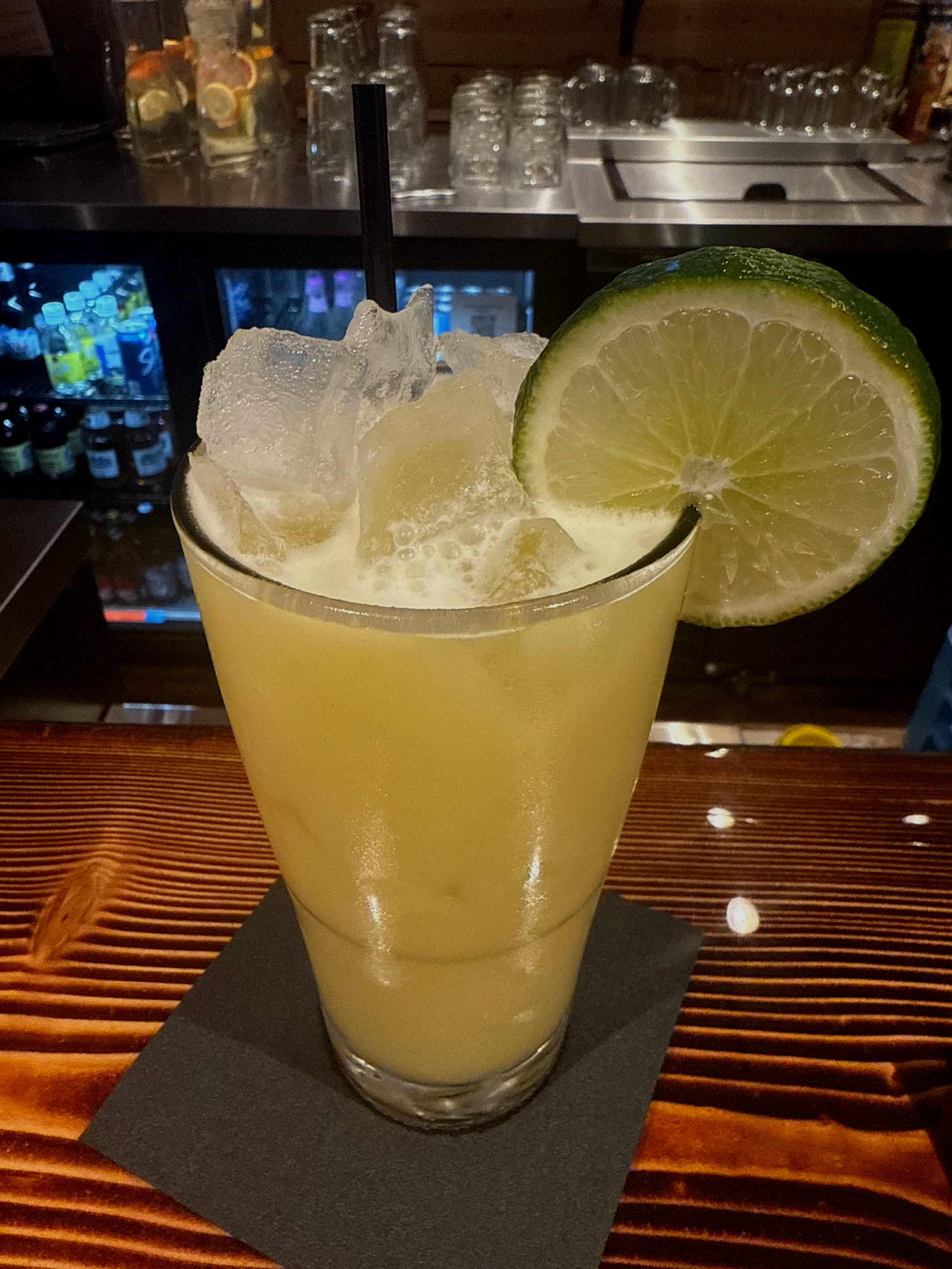 Cocktail in a tall glass on a bar, with a lime slice garnish.