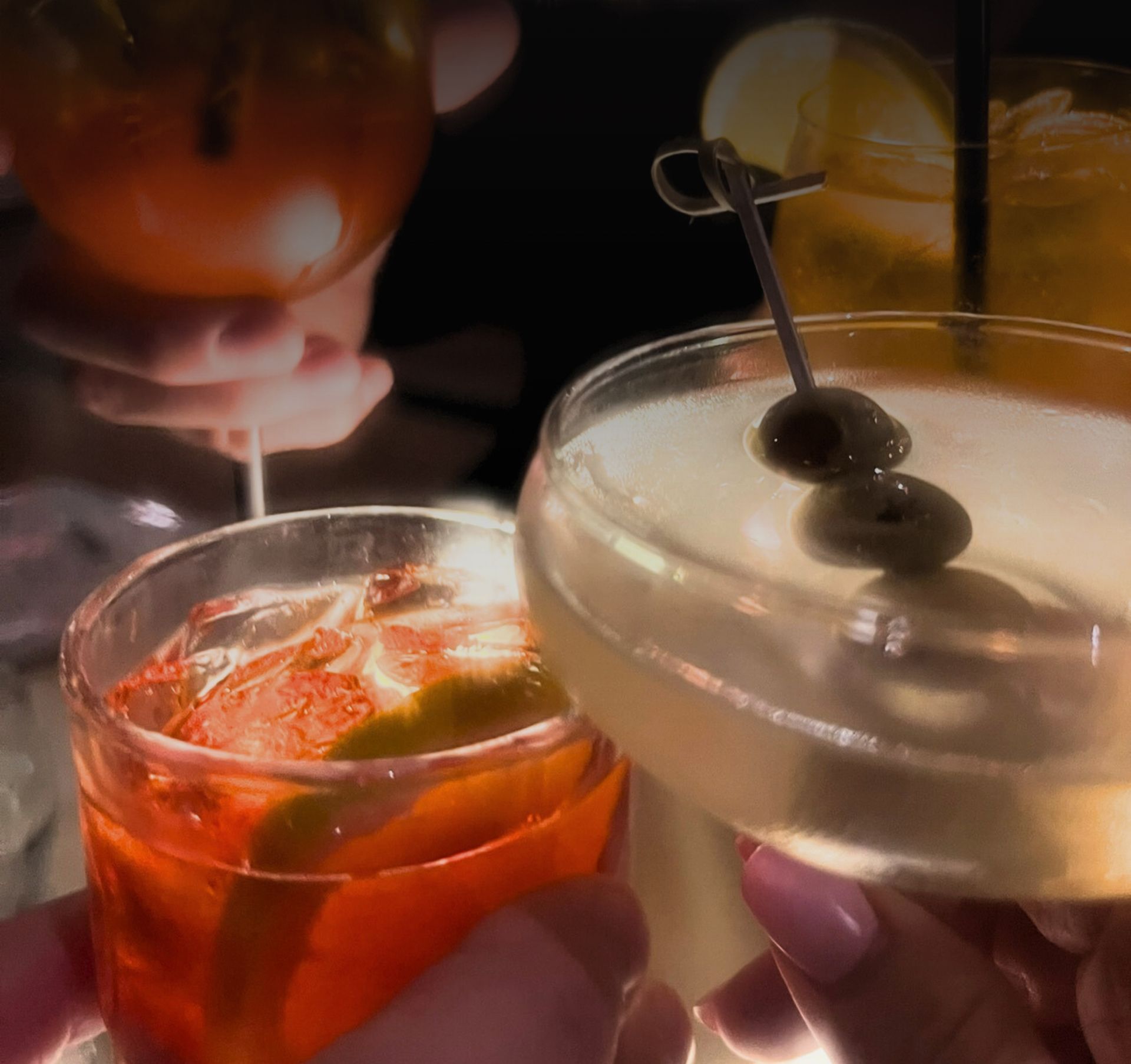 Two cocktails clinked together in warm, dim light, one orange and one pale with a cherry garnish.