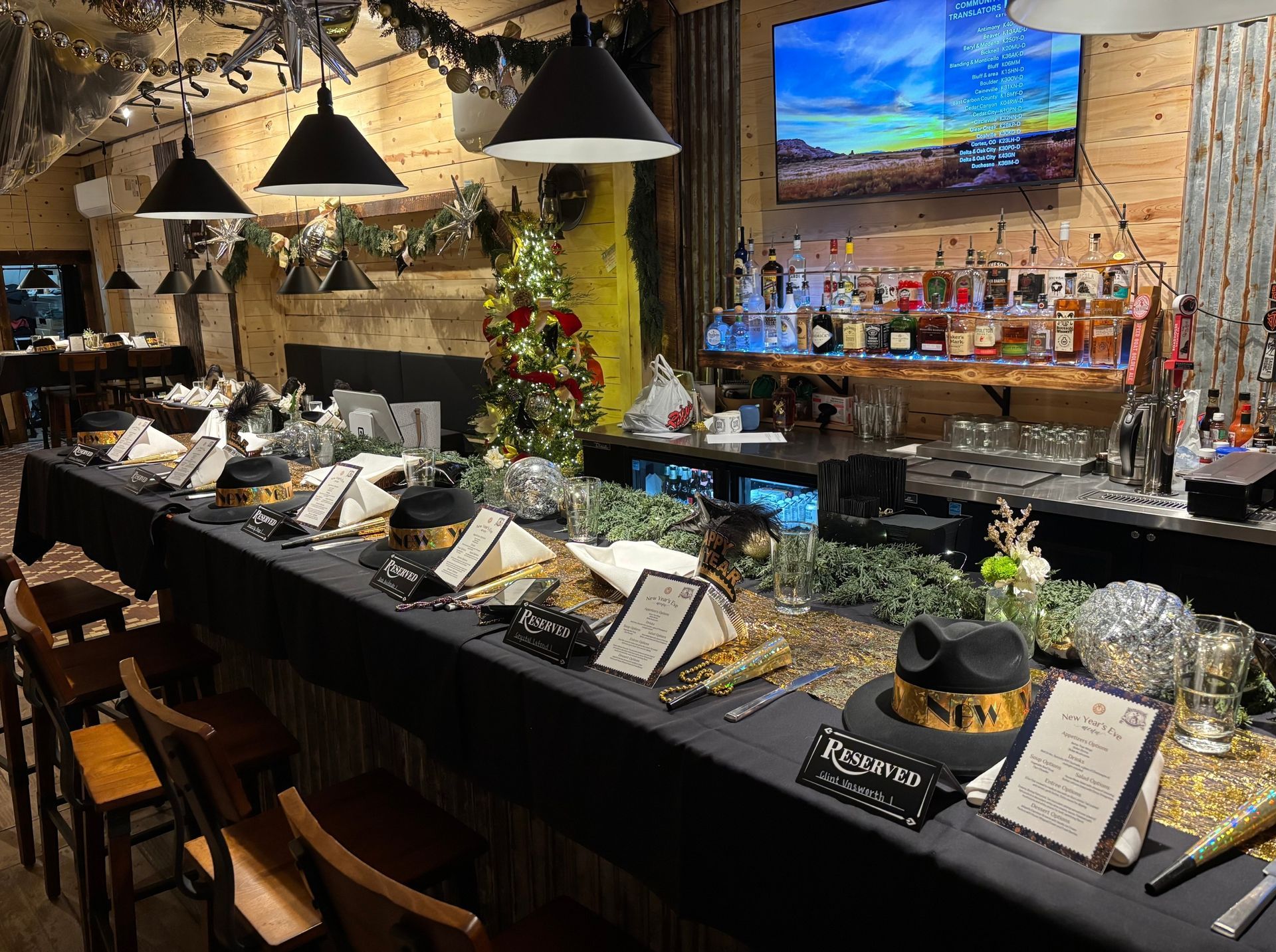Dimly lit buffet counter with labeled dishes, hanging lamps, plants, and a TV in a rustic restaurant interior