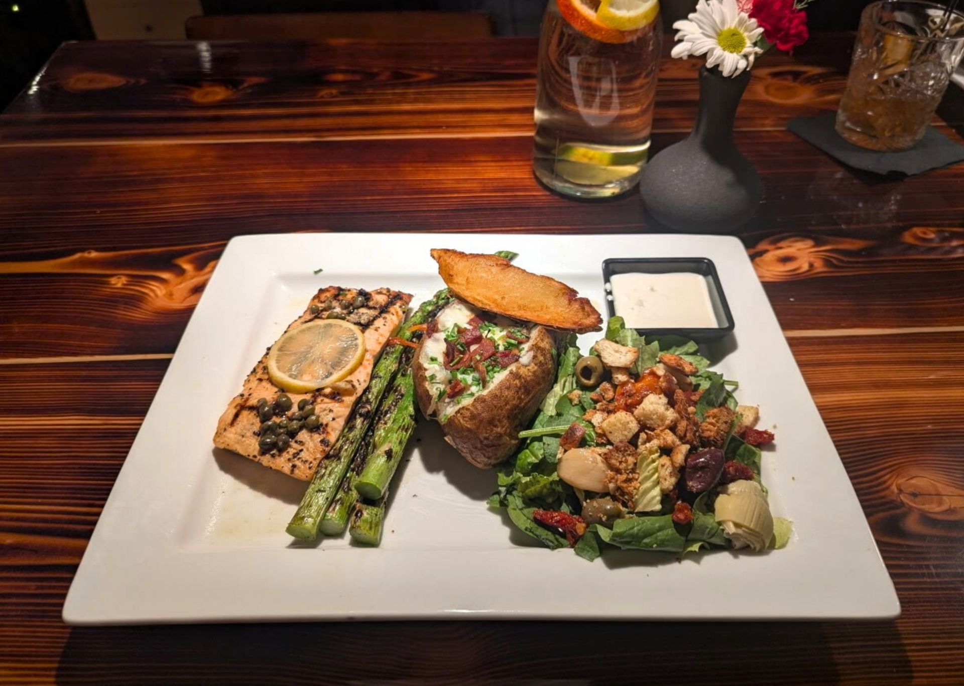 Plated gourmet fish and asparagus meal on a white square plate on a wooden table, with a drink nearby